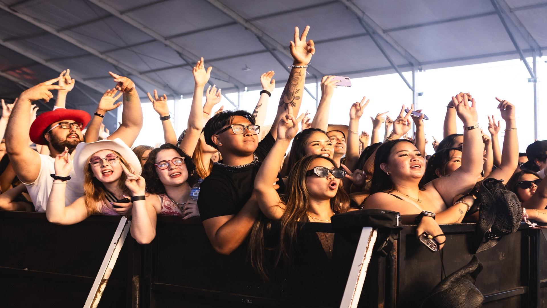 Concert crowd stands behind barrier, raising hands and phones under large tent structure.