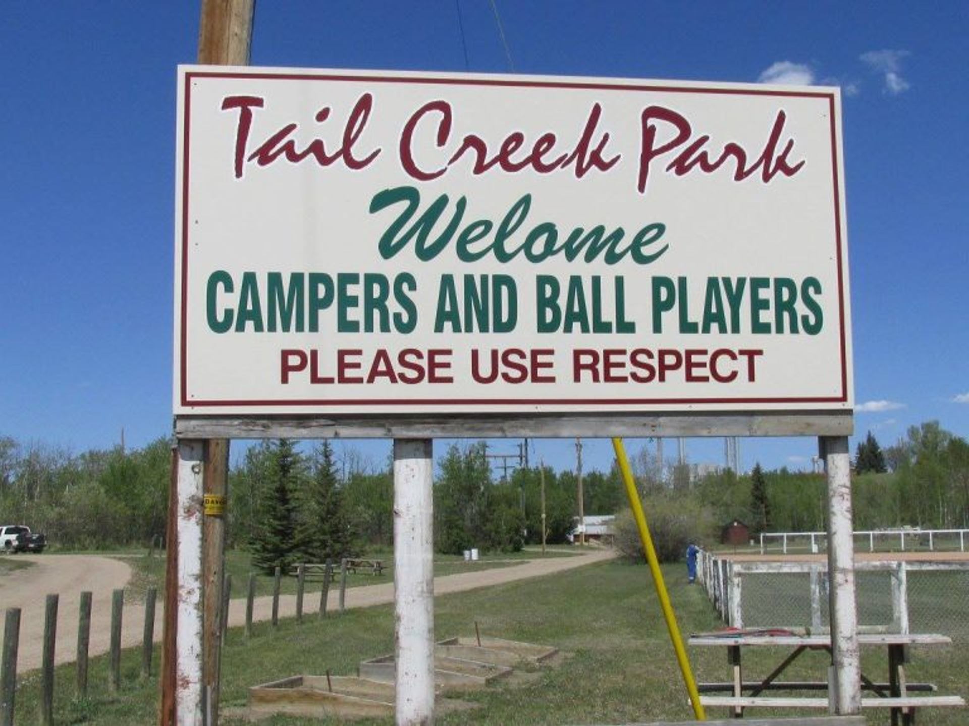 A Tail Creek Park welcome sign stands beside picnic tables and a rural park area.