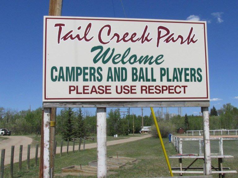 A Tail Creek Park welcome sign stands beside picnic tables and a rural park area.
