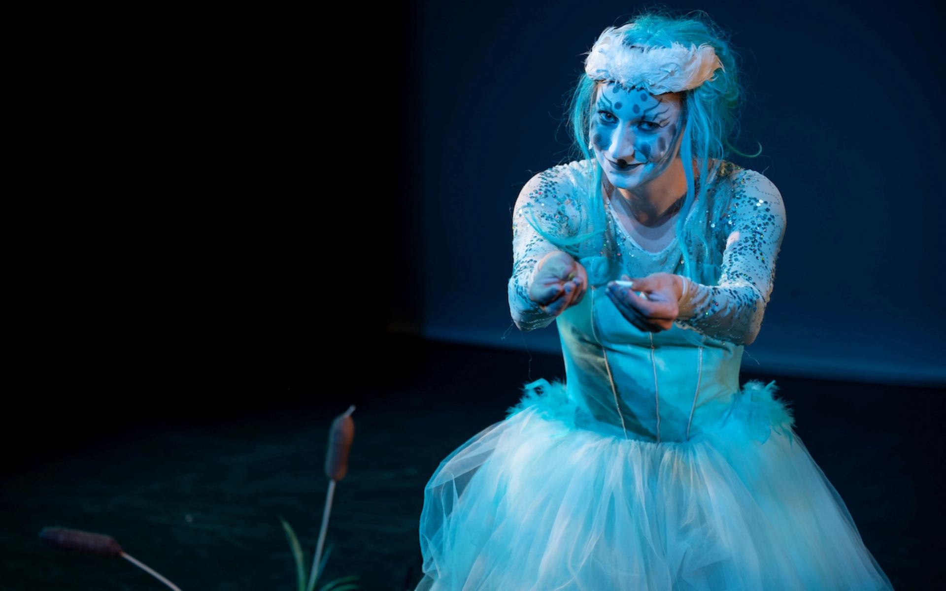 A performer in a fantastical blue costume with spotted face paint and blue hair holds a small object, standing near cattails on a dark stage.