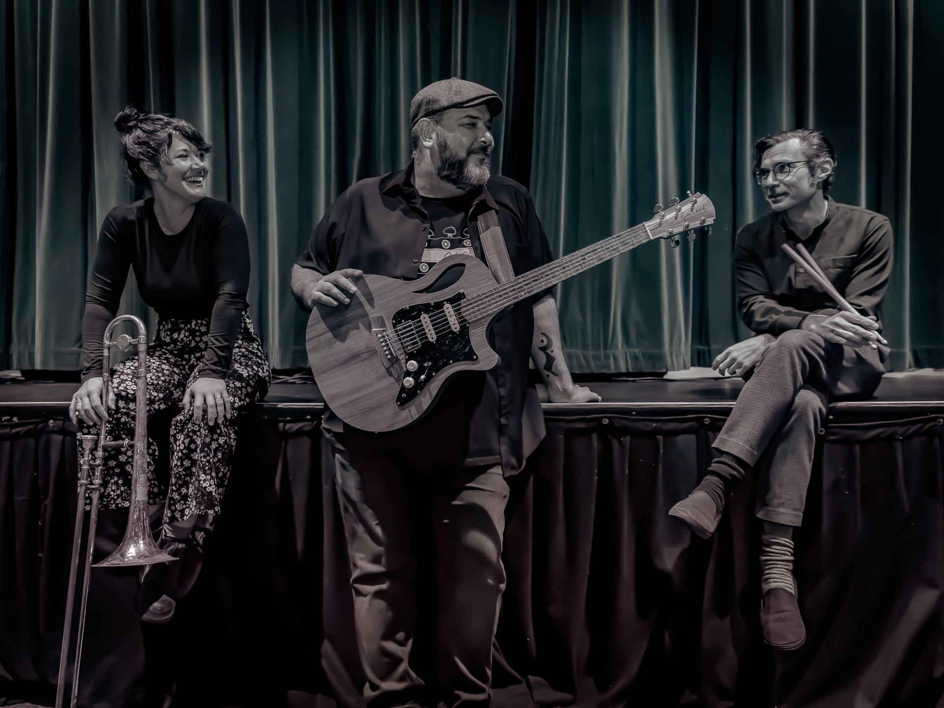 Three musicians with trombone, guitar and drumsticks sit on stage edge before curtains.