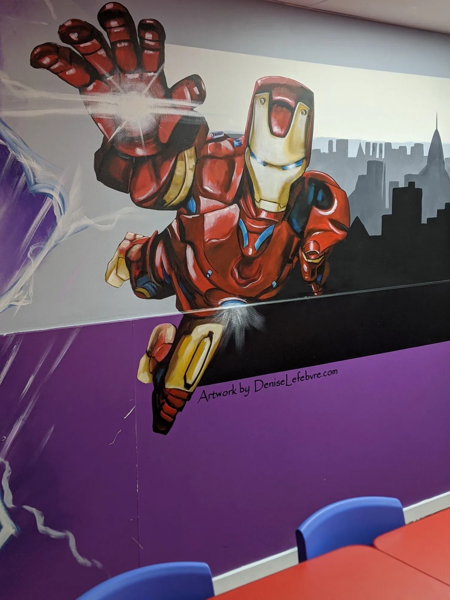 Wall mural of Iron Man flying forward, city skyline silhouette, colourful chairs below.