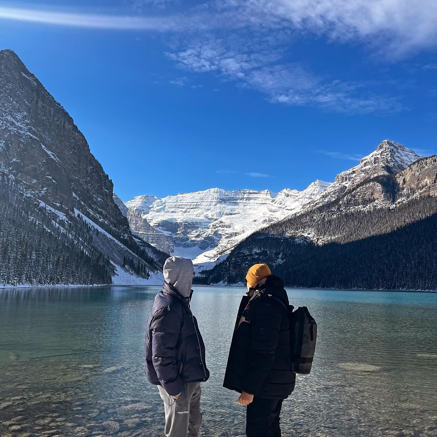 Two people stand by a turquoise lake, looking at snow-capped mountains under a bright blue sky.