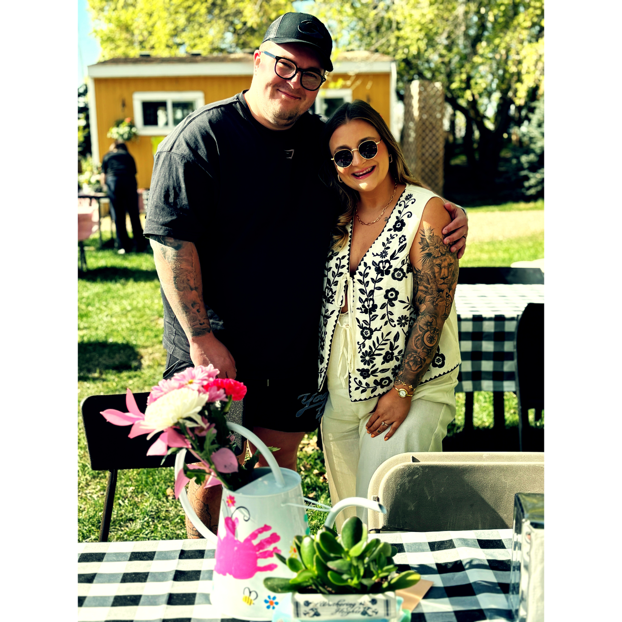 Two people with tattoos stand together outdoors behind picnic tables and flower decorations.