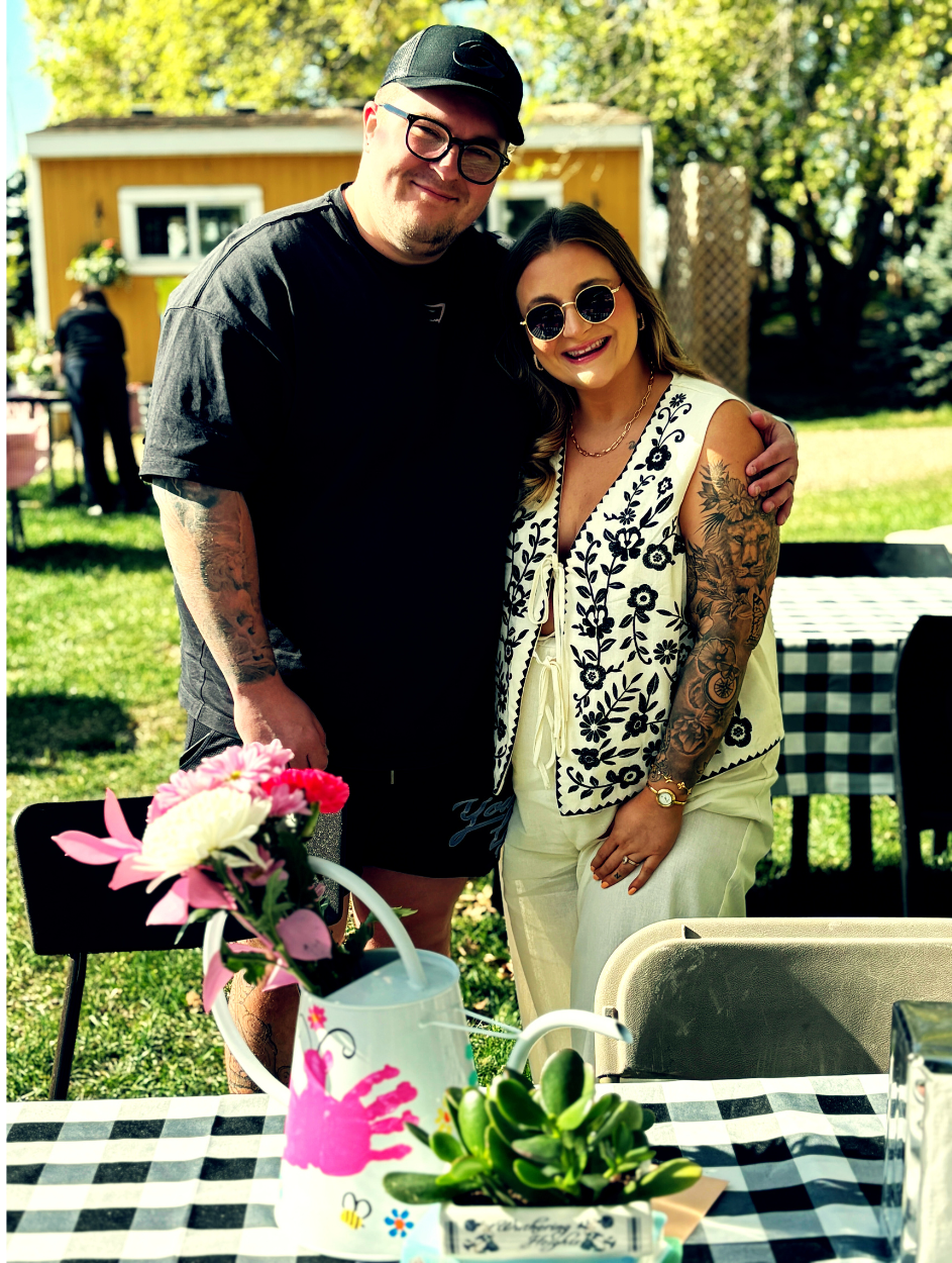Two people with tattoos stand together outdoors behind picnic tables and flower decorations.