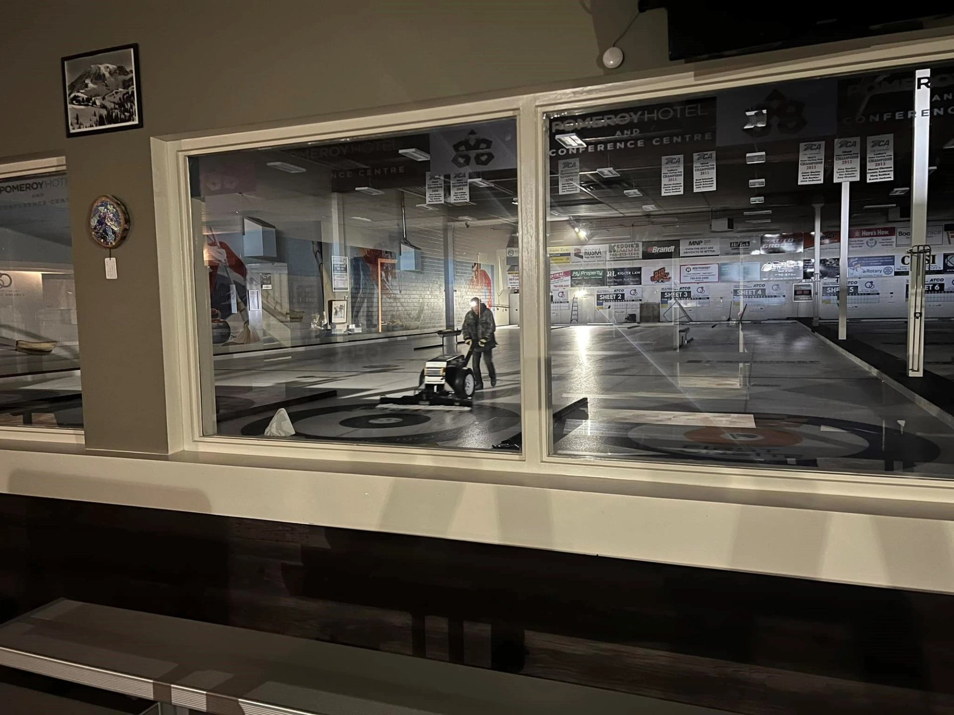 Window view of curling rink as person pushes ice maintenance machine.
