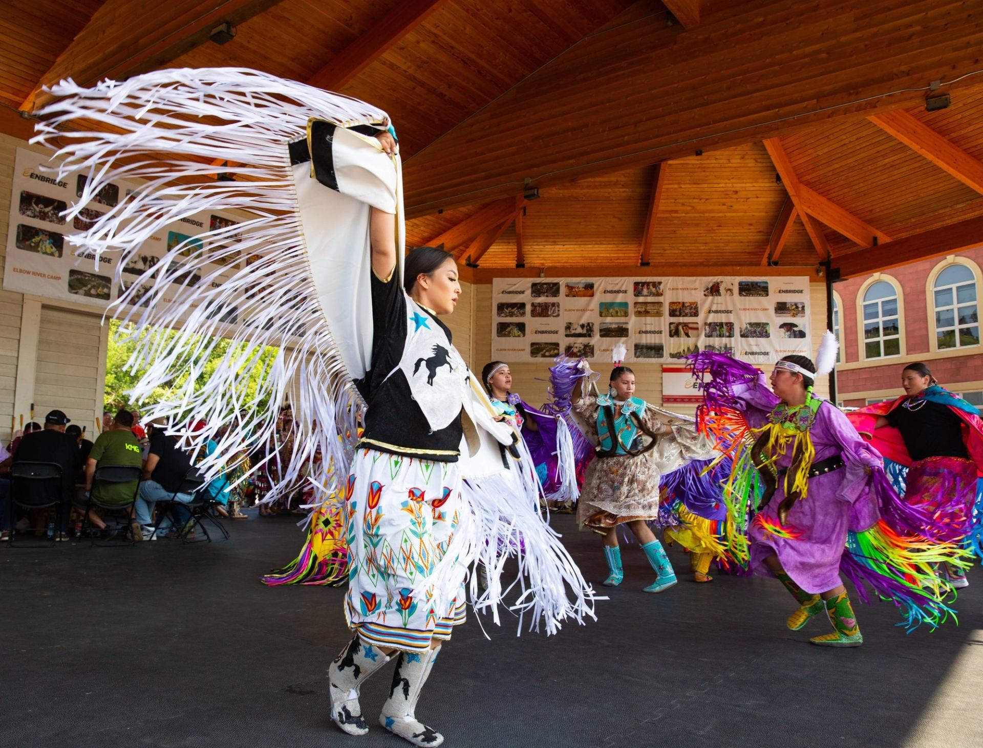 Dancers in colourful regalia perform under a wooden pavilion before seated spectators.