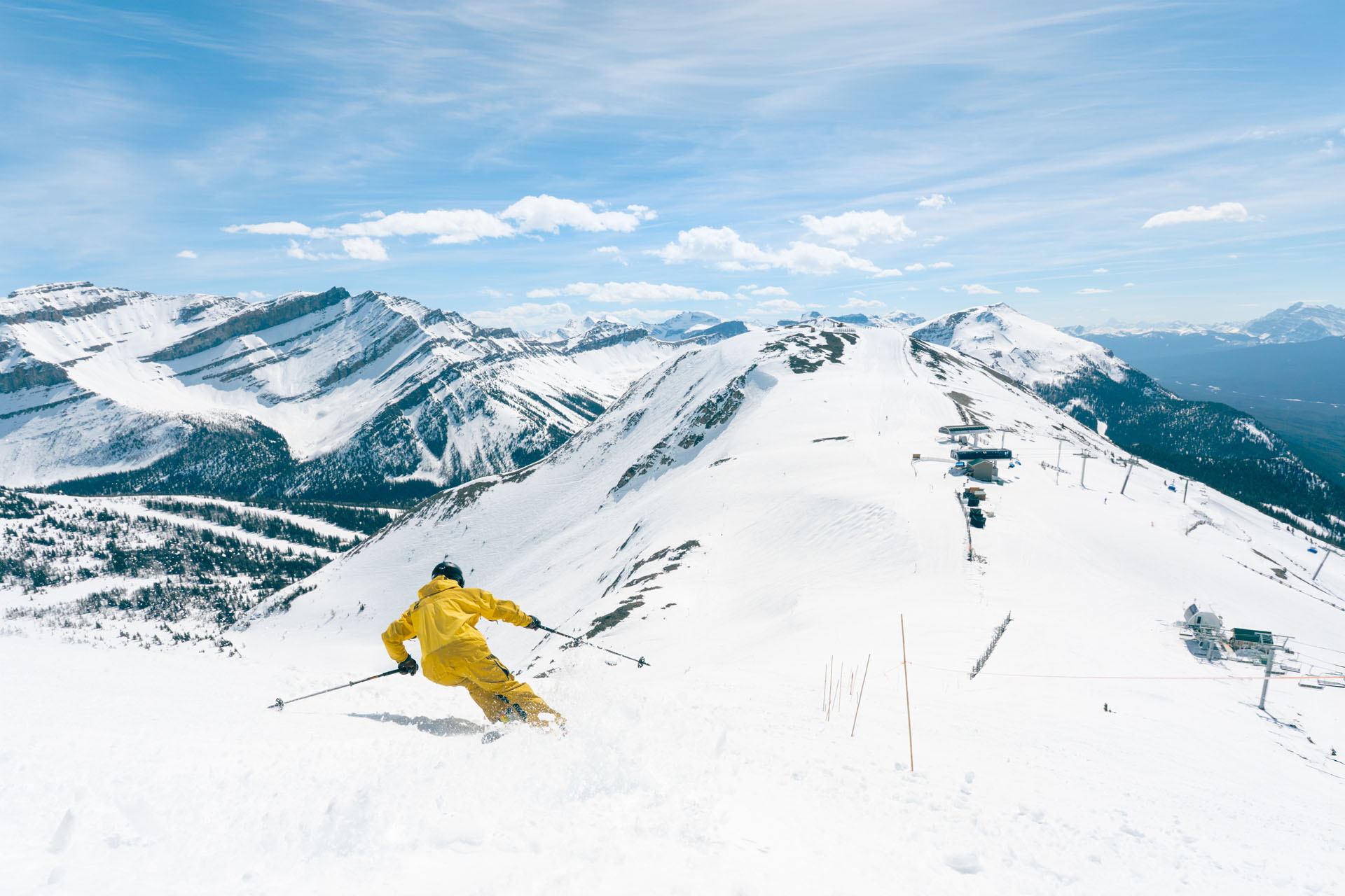 Skier in yellow suit descends snowy mountain slope with chairlifts and distant peaks.