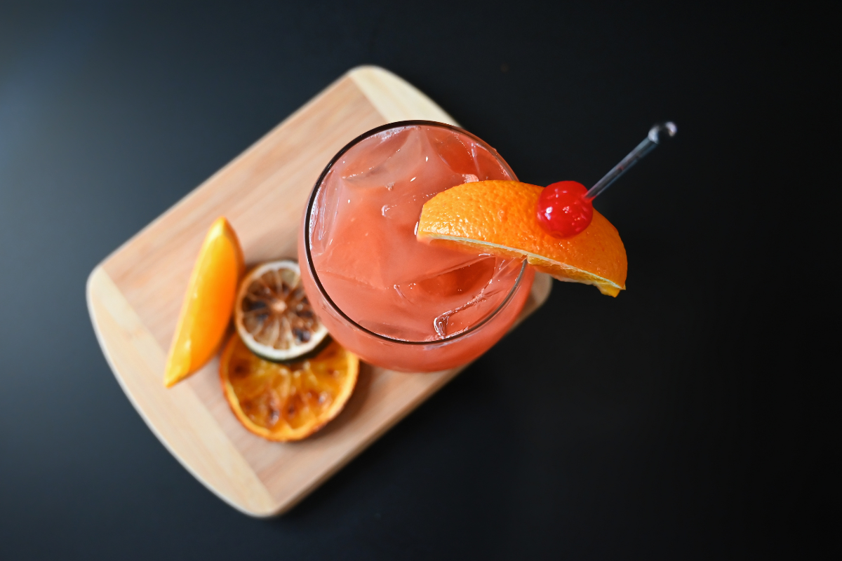 Pink-orange cocktail with cherry and orange garnish on a wooden board.