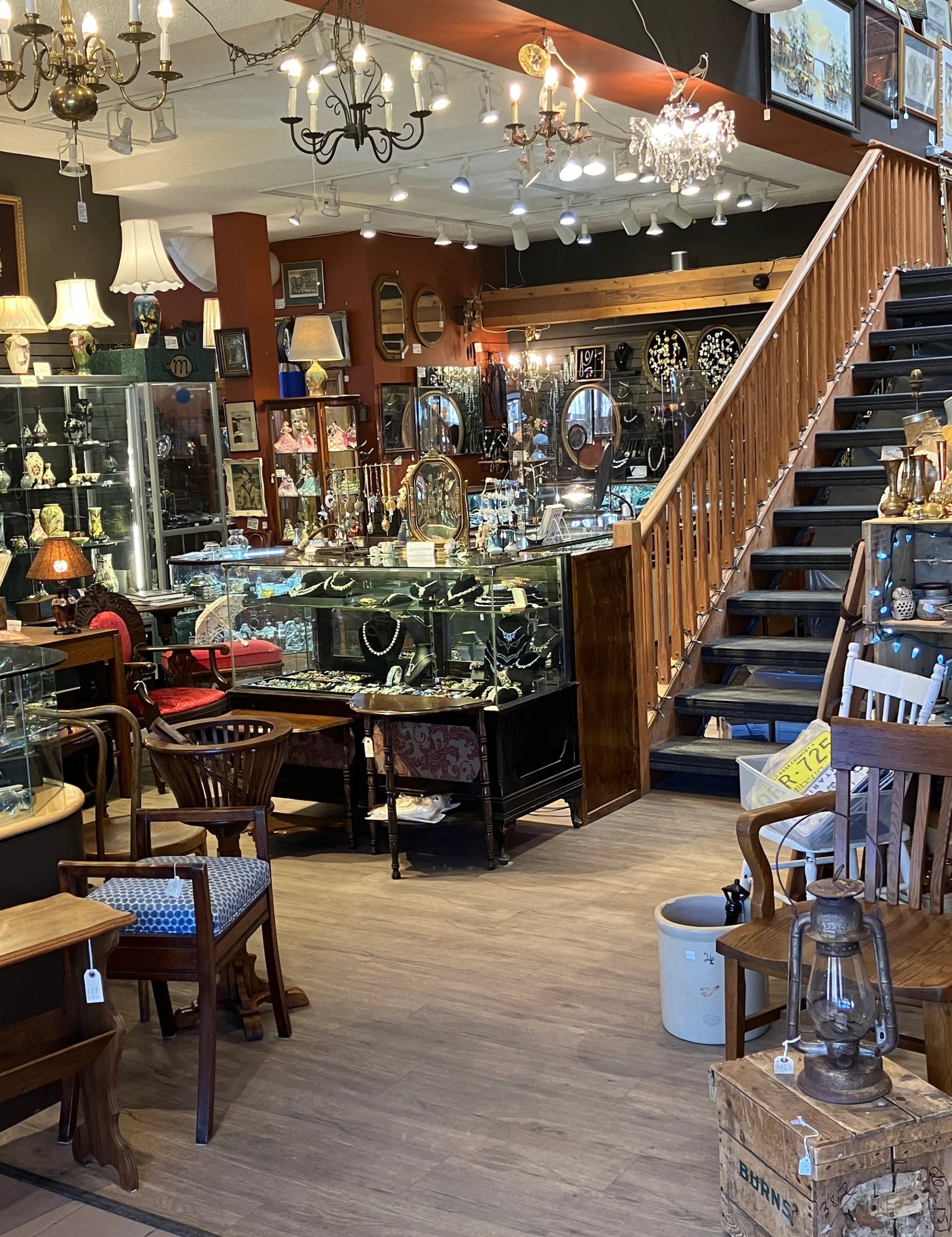 Antique shop interior with glass jewellery cases, wooden furniture and staircase under hanging lights.