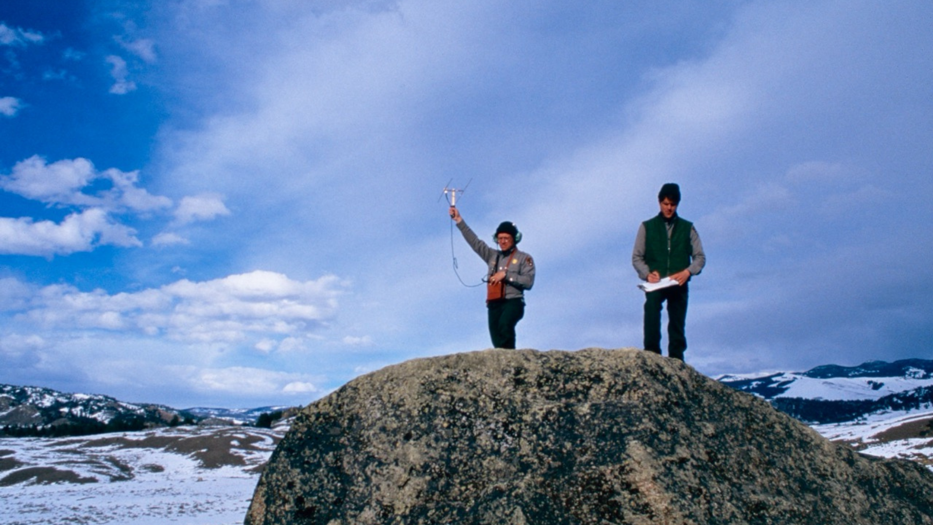 Two people stand on a rocky hill, using antenna equipment and writing notes.