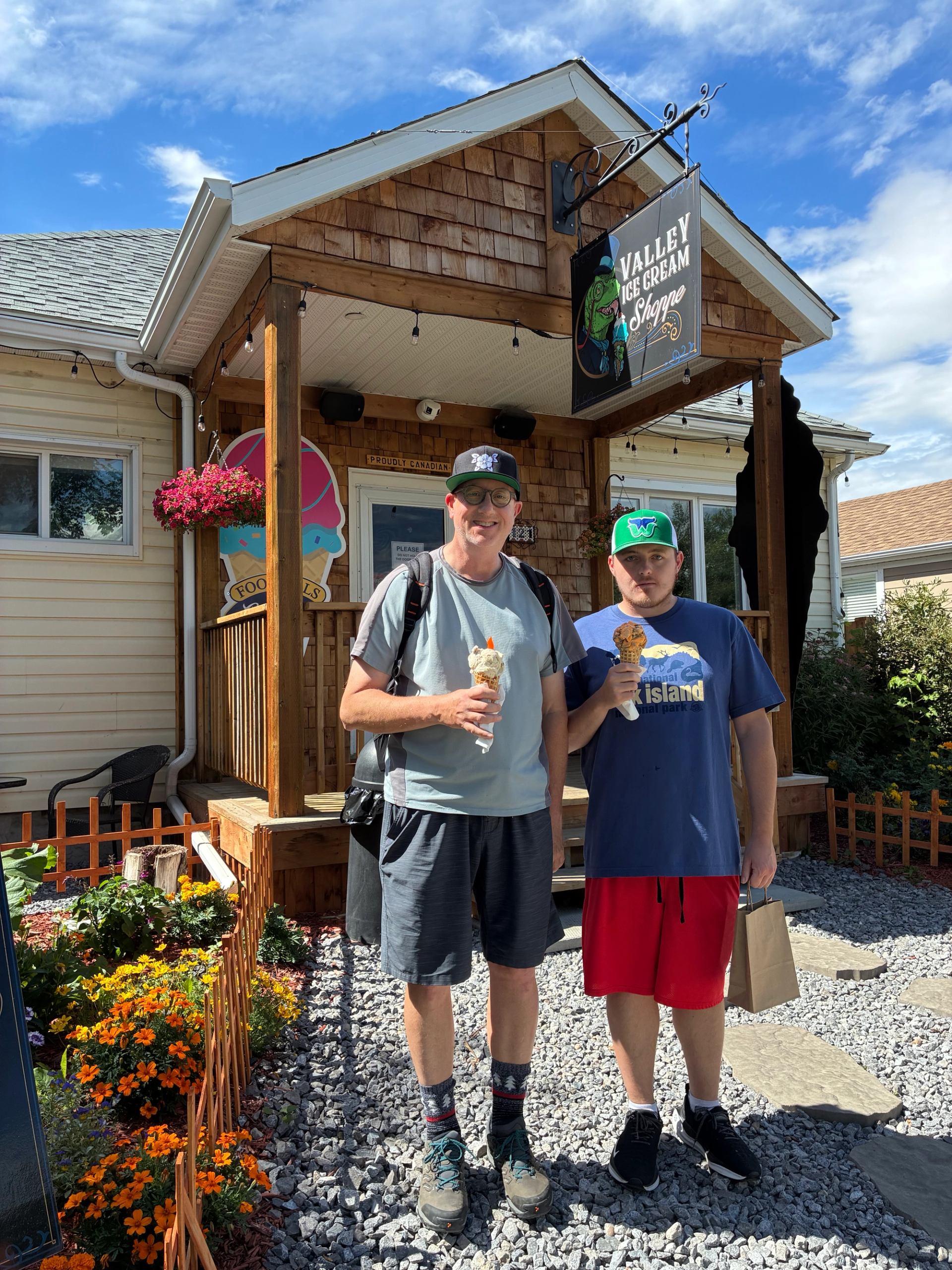 Two visitors stand outside a quaint ice cream shop holding cones on a sunny day.