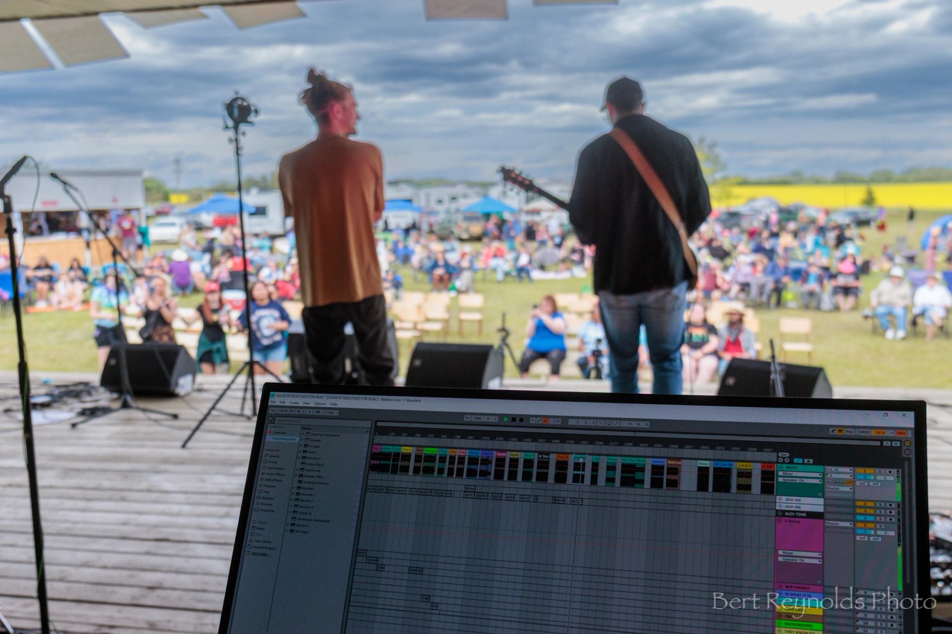 Outdoor concert stage with two performers, laptop audio software, and seated crowd in field.