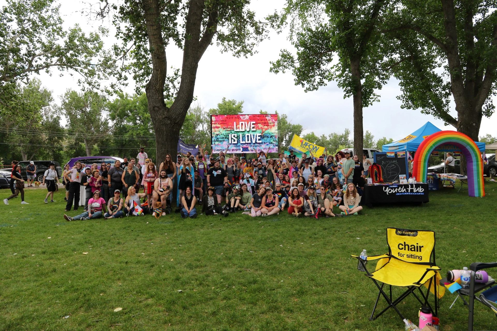 Large group at outdoor Pride event with Love Is Love sign and rainbow decorations.