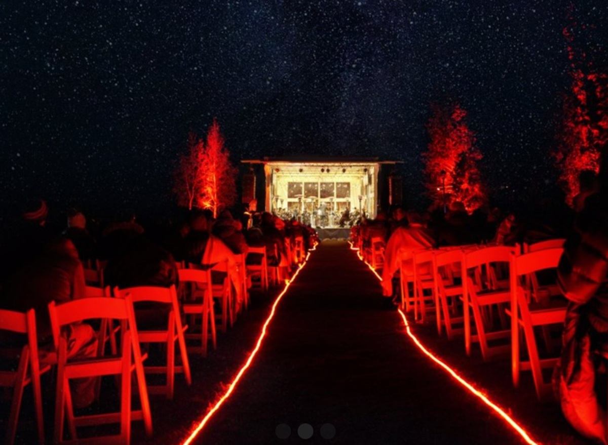 Outdoor audience sits on red-lit chairs facing illuminated stage under starry night sky.