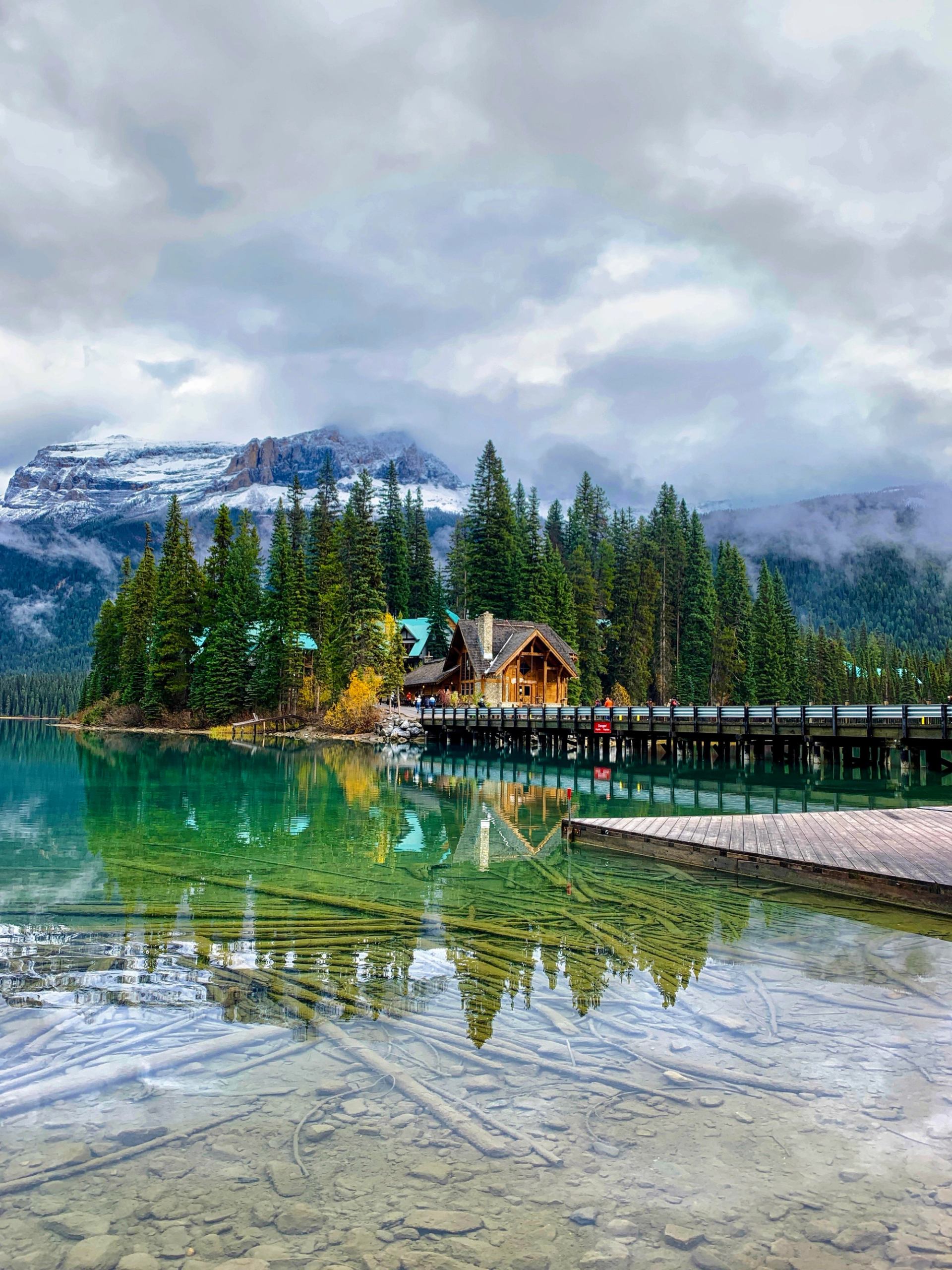 Turquoise lake reflects a wooden lodge, evergreen forest and snow-covered mountains.