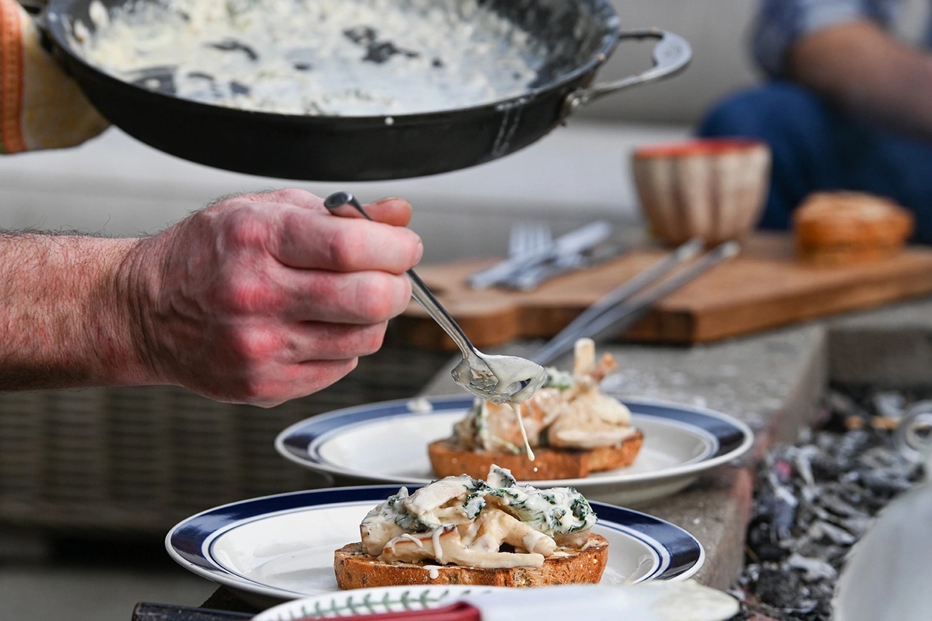 Cook’s hand spooning creamy sauce over toast with mushrooms on outdoor plates.