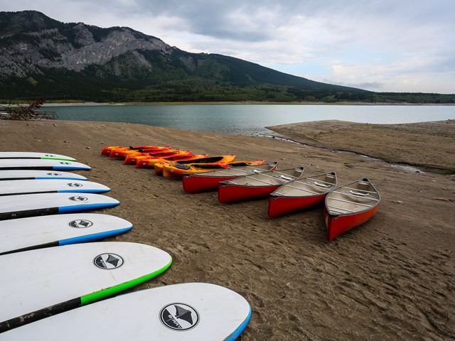 Paddleboards, kayaks, and canoes lined up on a beach with mountains in the background.