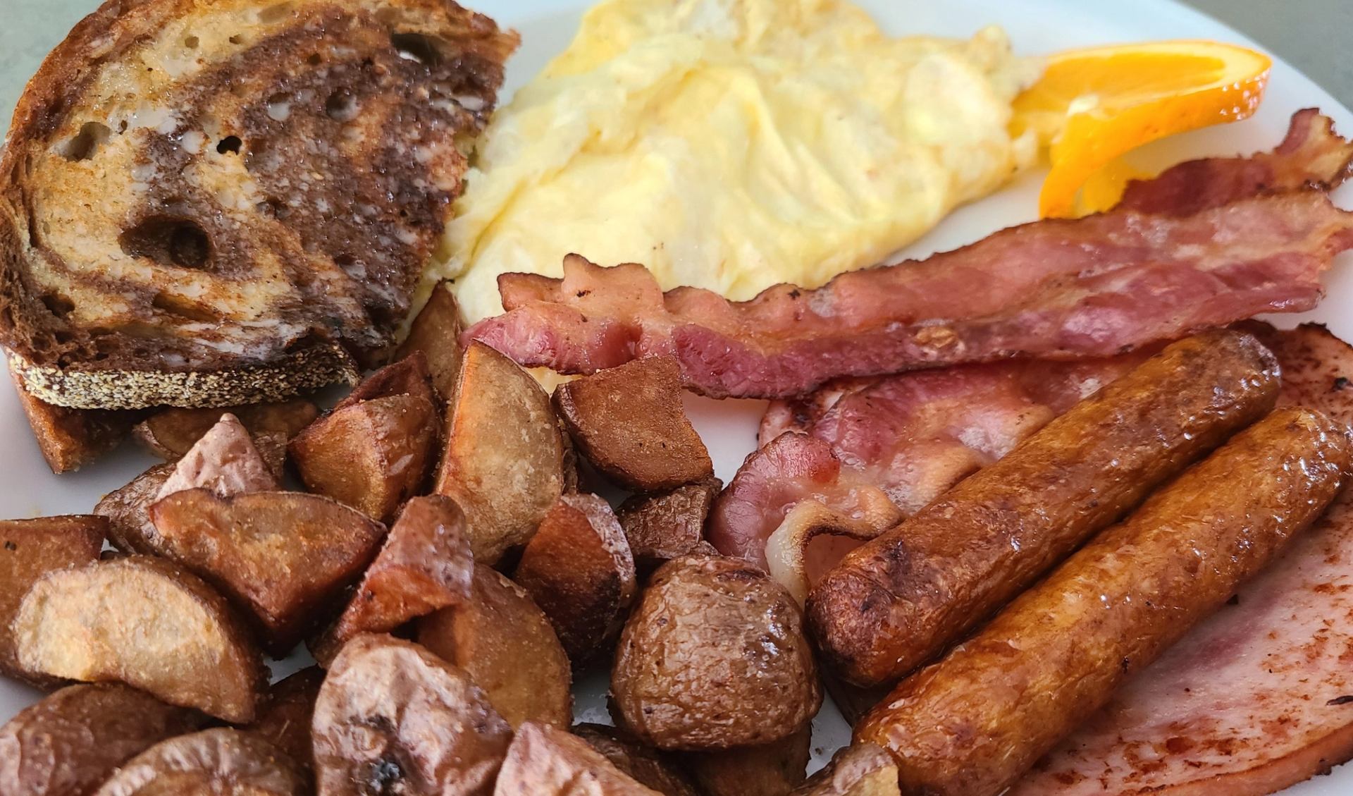 A hearty breakfast plate with scrambled eggs, bacon, sausage, ham, home fries, and toasted bread.
