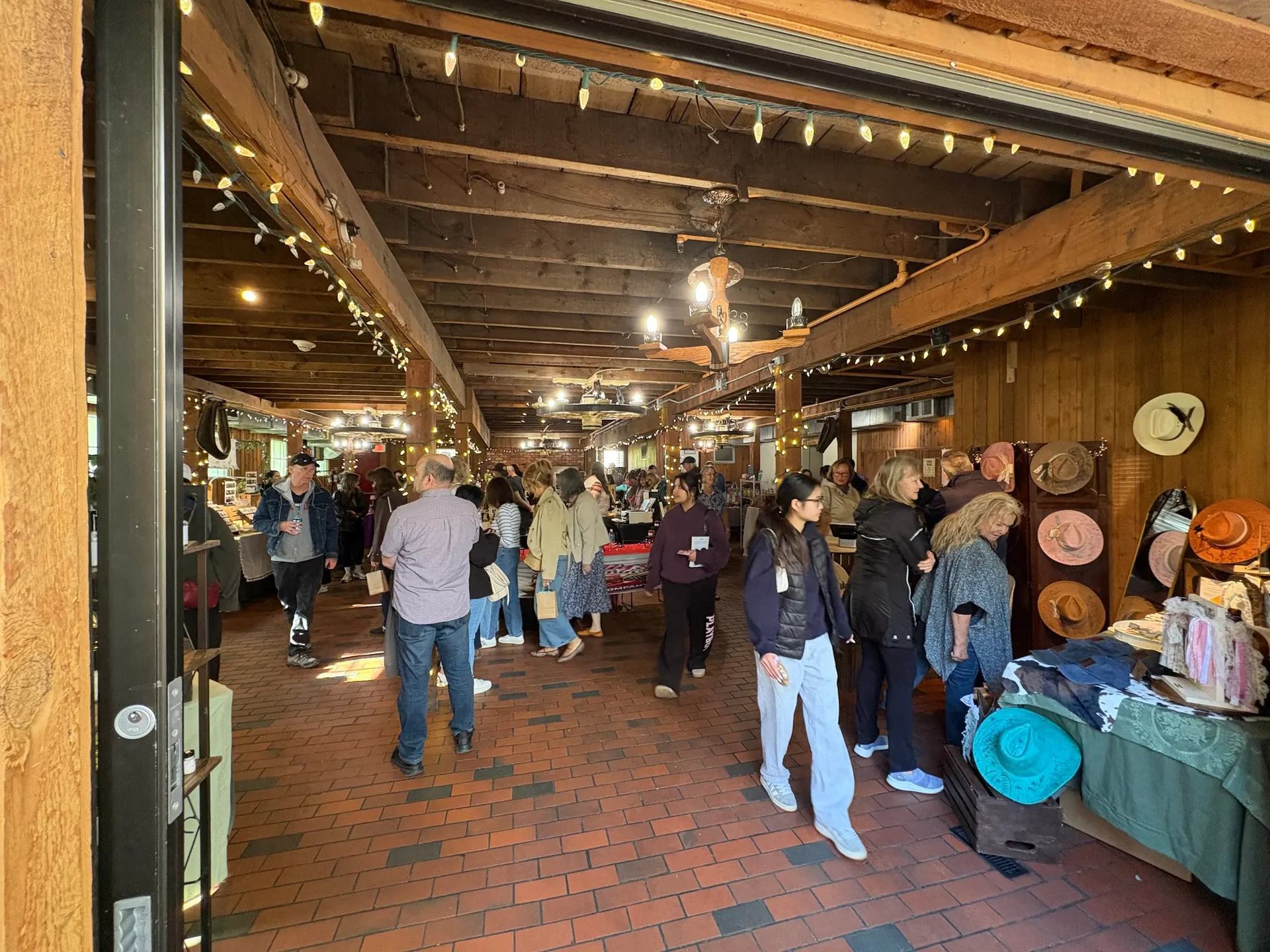 Indoor craft market with shoppers browsing hat displays and vendor tables under string lights.