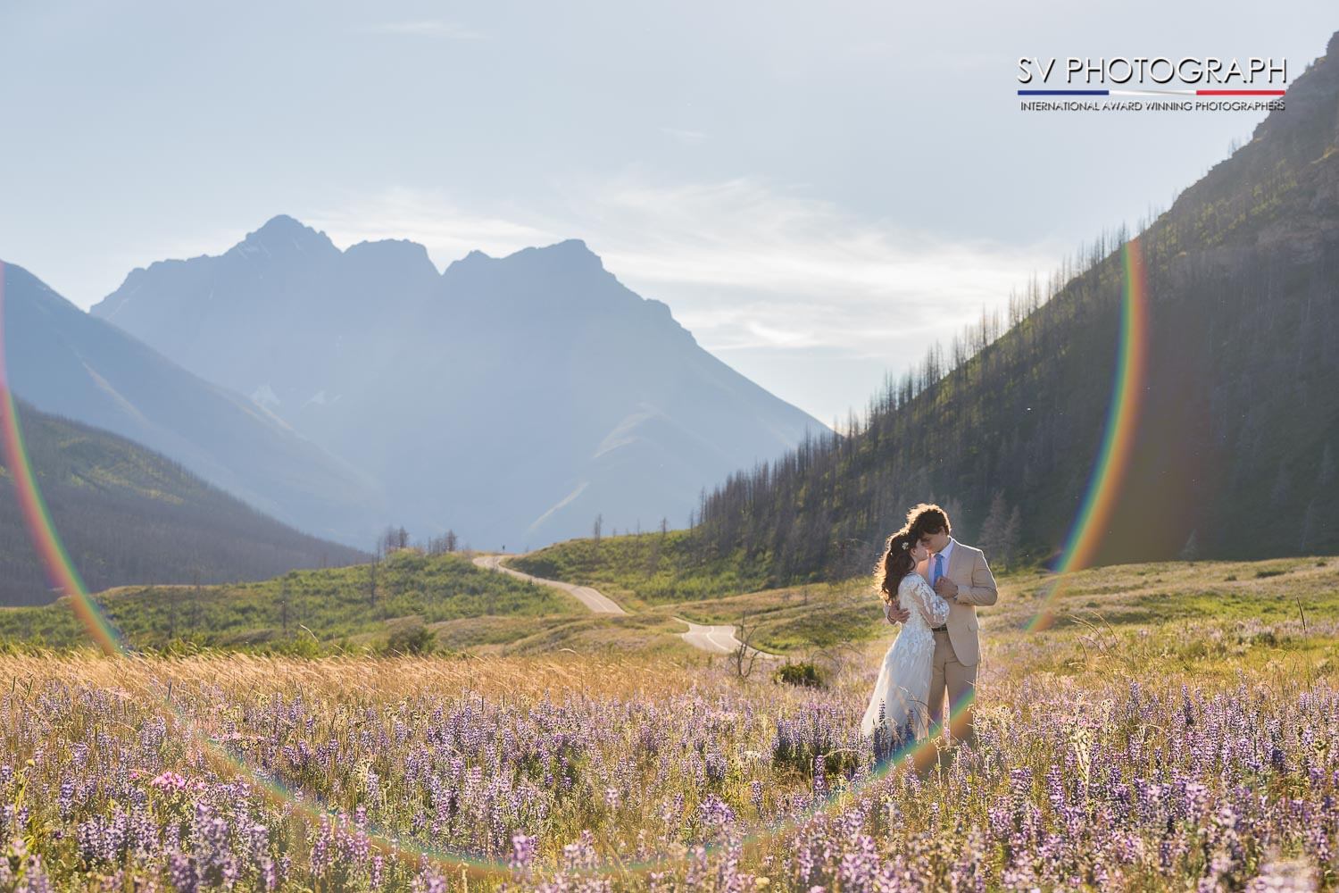 Couple embraces in a field of purple wildflowers under a bright sky, mountains behind and a rainbow halo around them, creating a romantic, serene atmosphere.