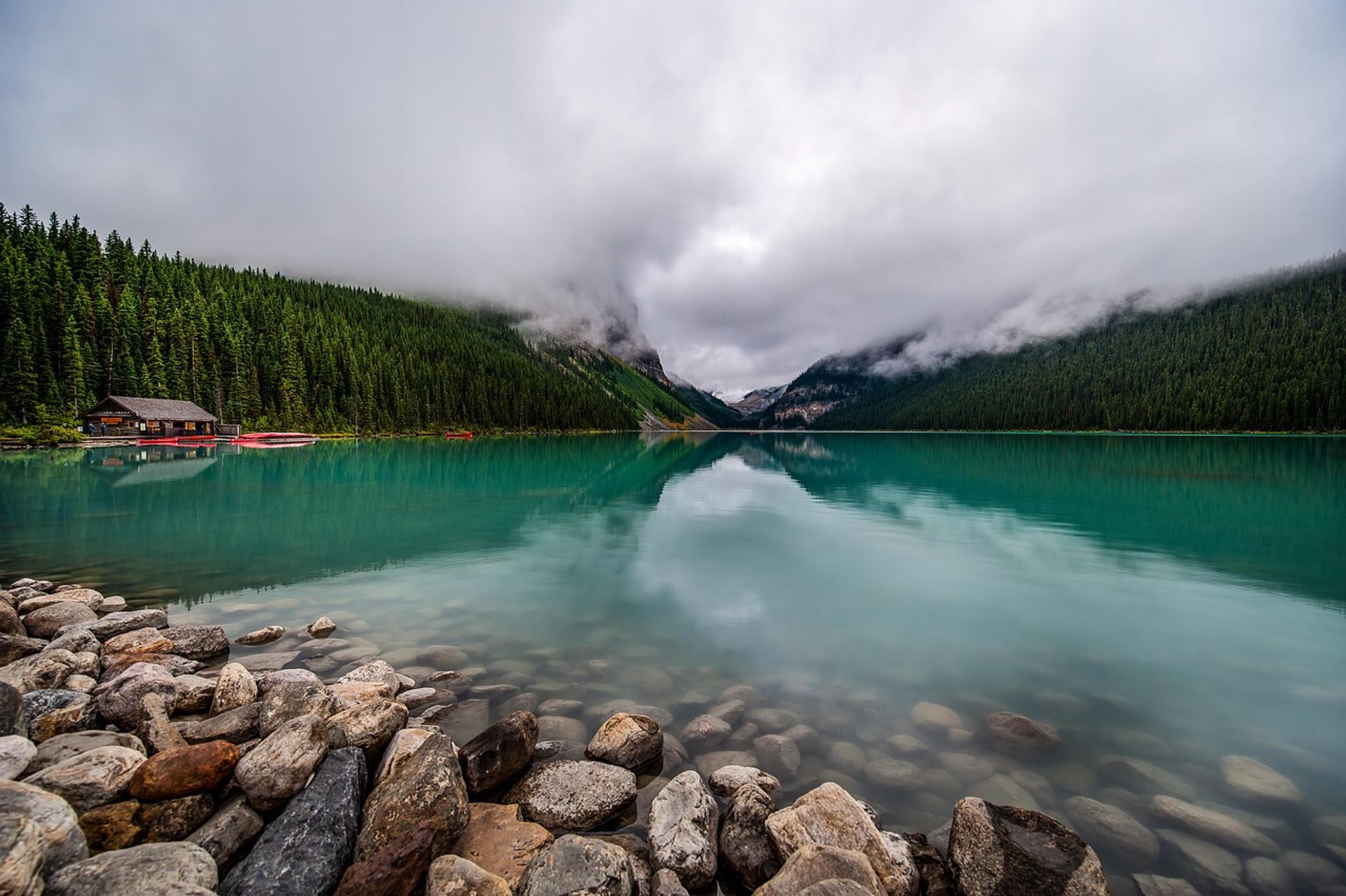 Rocky lakeshore with turquoise water, forested hills, low clouds and small wooden cabin.