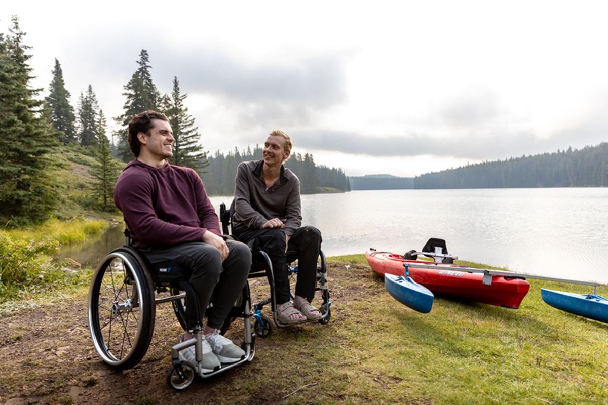 Two wheelchair users sit by a forest lake beside an adaptive red kayak.