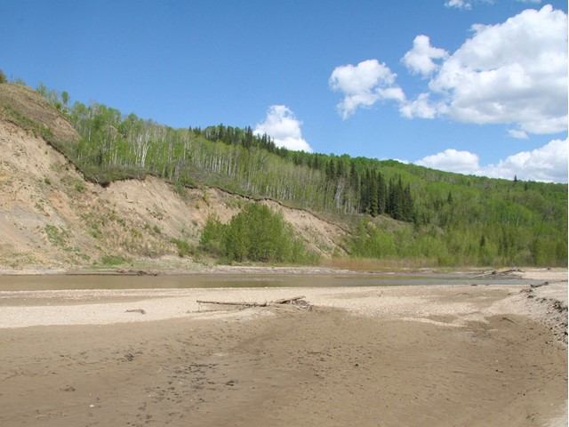 A wide river with sandy and muddy banks, flanked by steep, forested hills under a blue, cloudy sky.