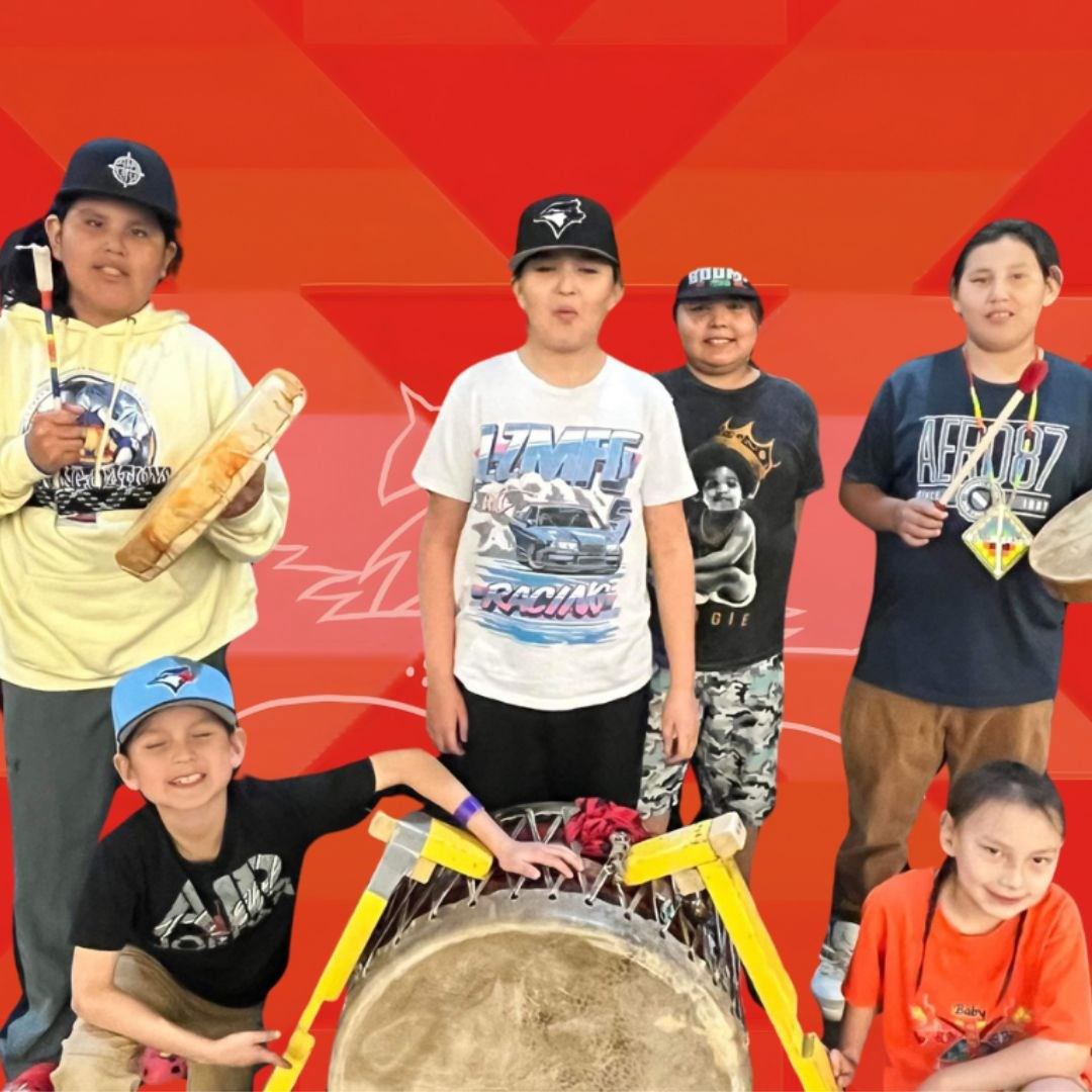 Group of children with drums and sticks posing against a bright red geometric backdrop.