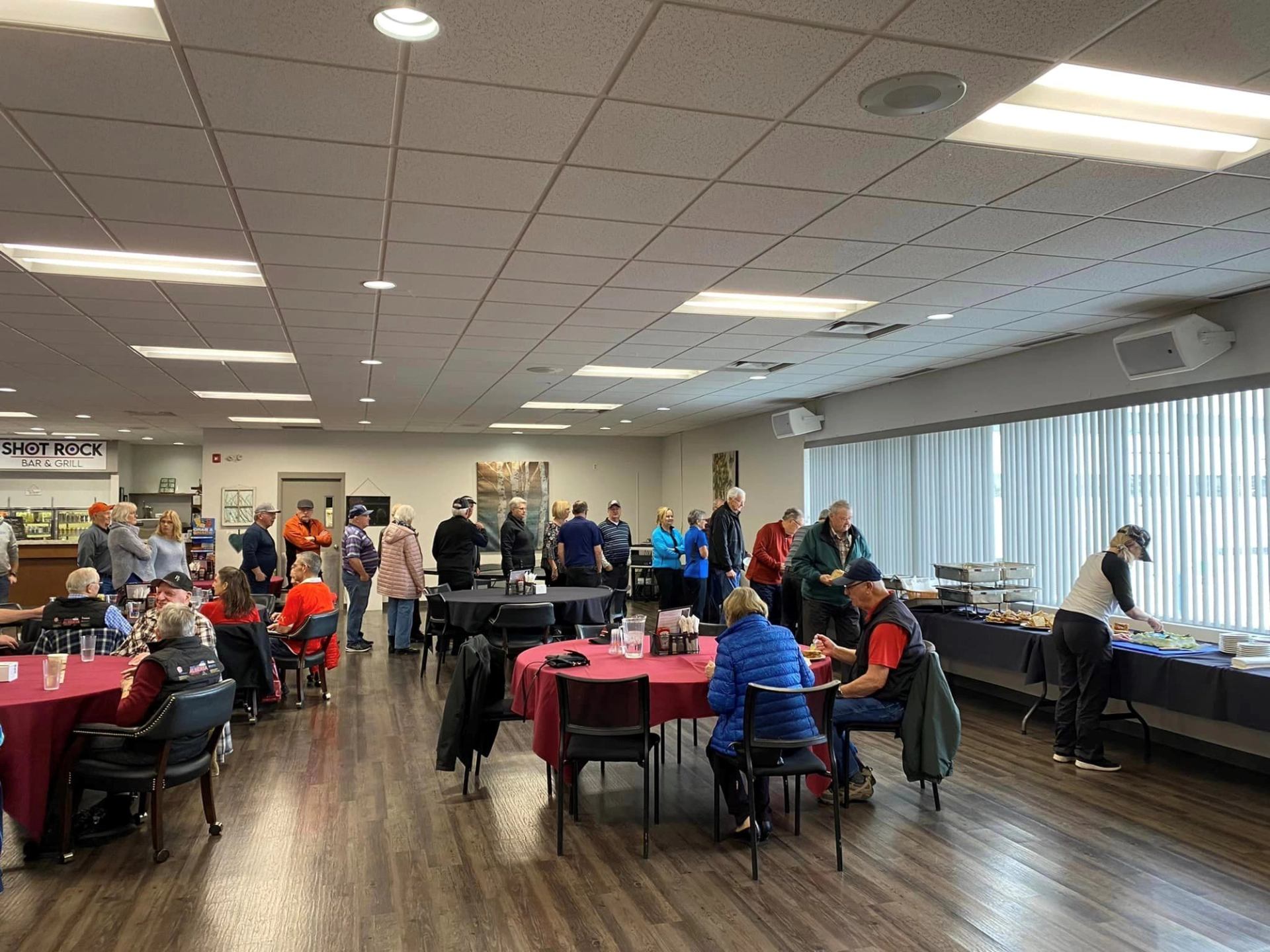 People sit at round tables and line up at buffet beside windows.