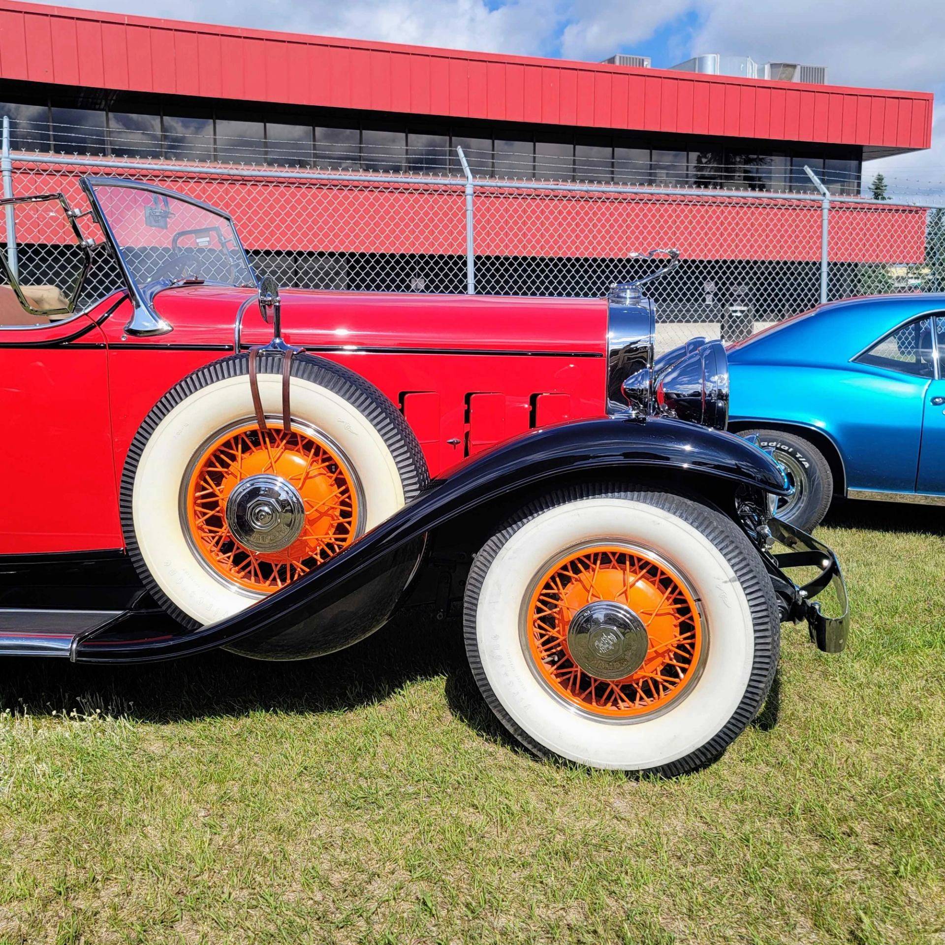 Vintage red convertible with orange-spoked whitewall tires parked beside blue car on grass.