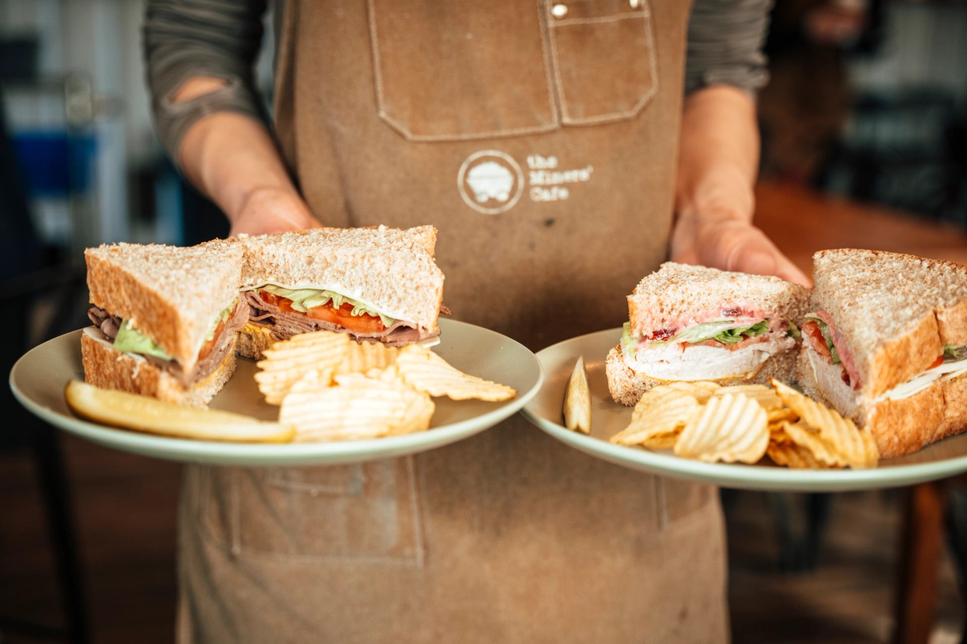 Staff member carrying two freshly made sandwiches with kettle chips on serving plates.