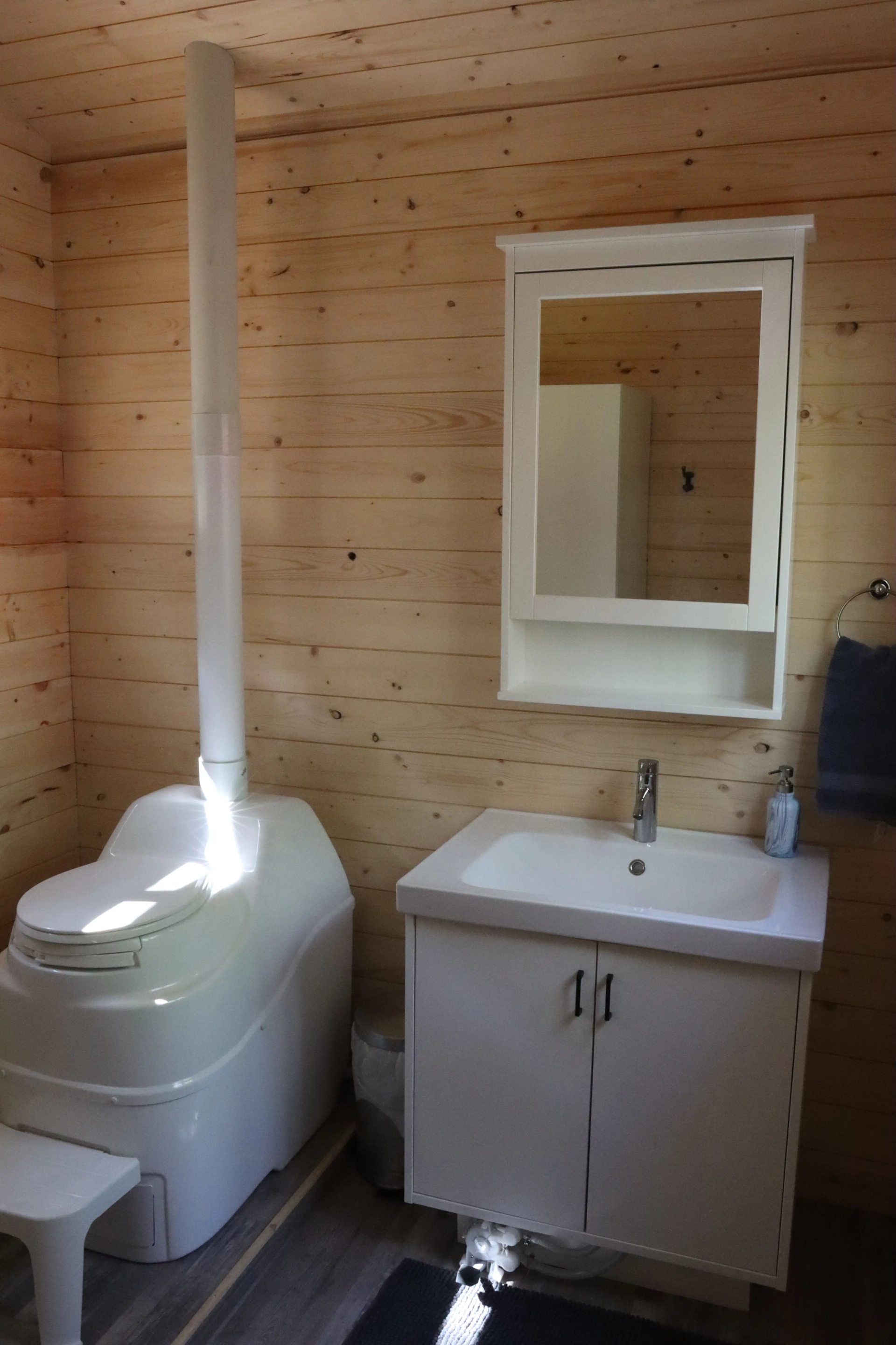 Small bathroom with composting toilet, sink, mirror, towel, and wood-paneled walls.