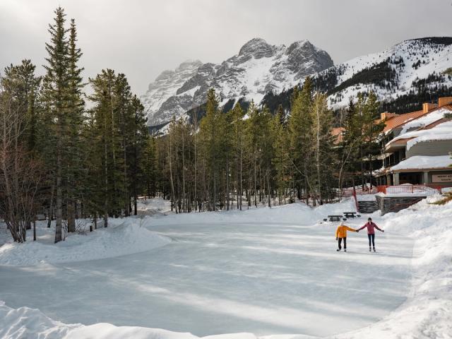 A couple holding hands and ice skating, surrounded by trees and mountains, on an outdoor rink at the Pomeroy Kananskis Mountain Lodge