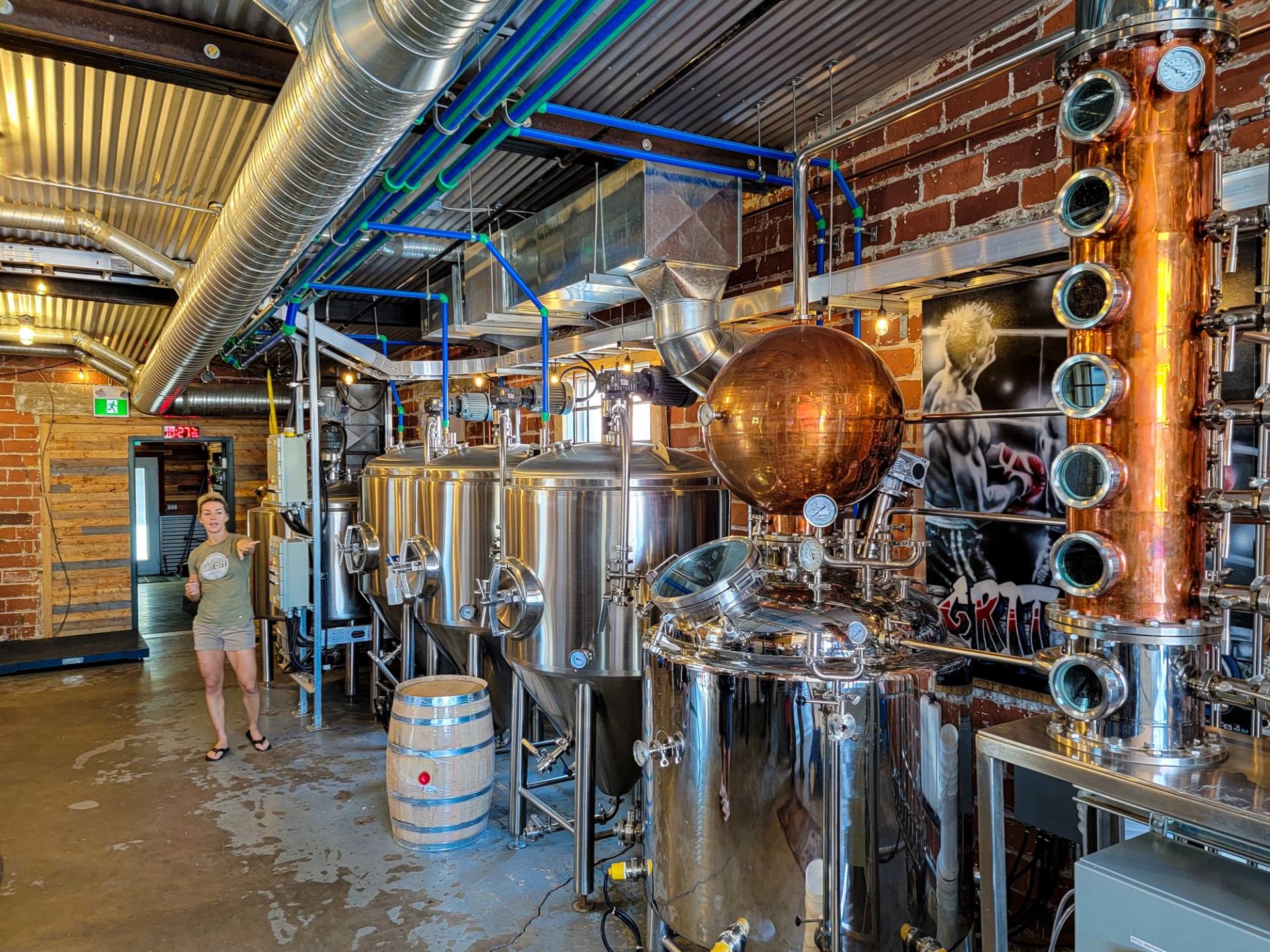 Guide stands by brewing tanks and copper still inside a craft distillery.