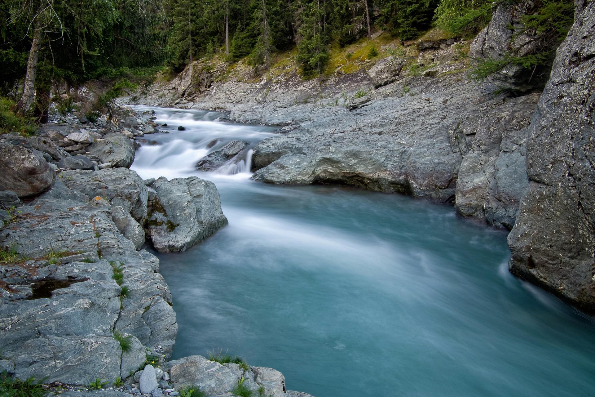 Turquoise river flows between smooth grey rocks below dense evergreen forest.