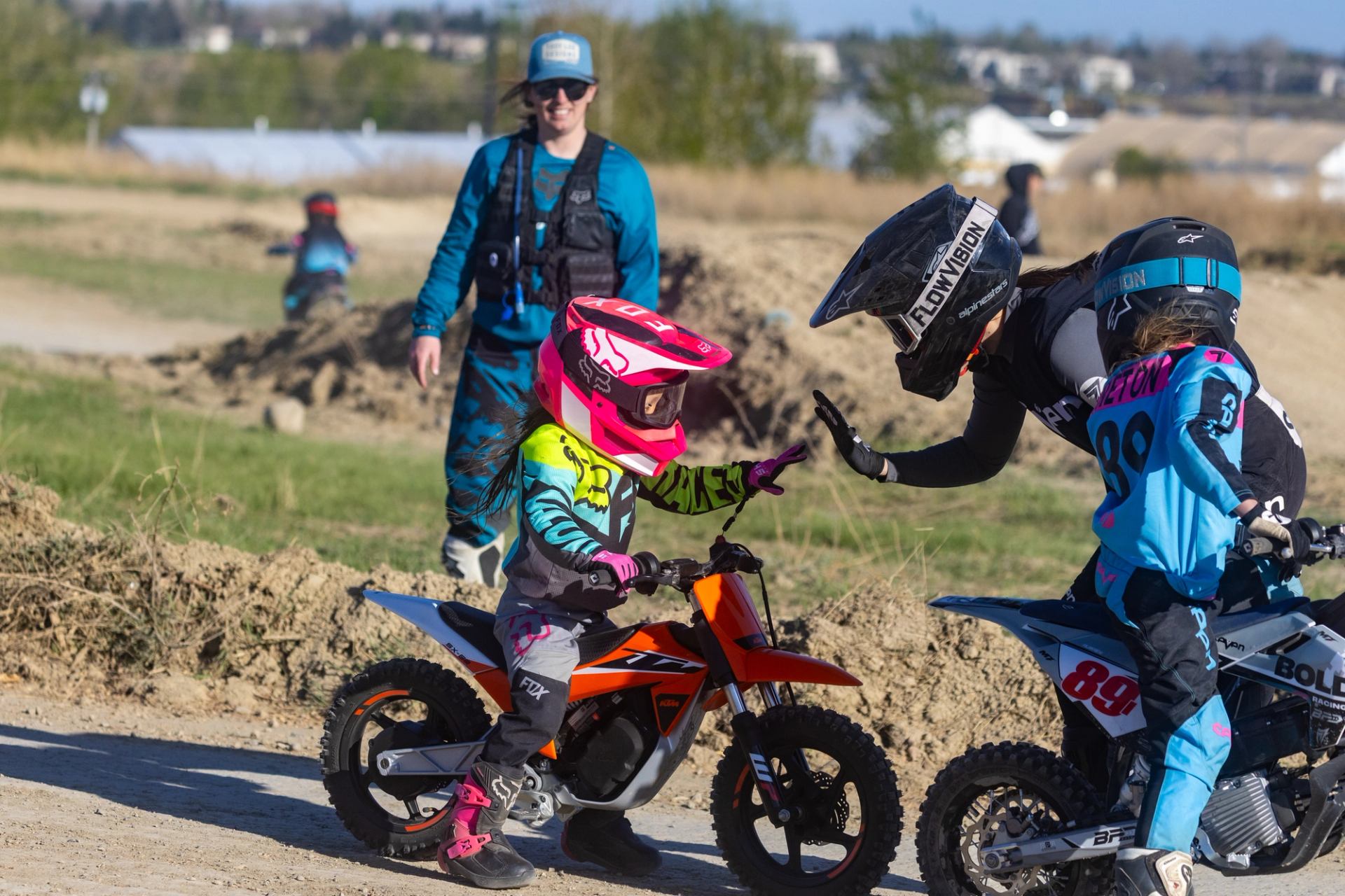 Two young motocross riders high-five on dirt bikes at outdoor track.