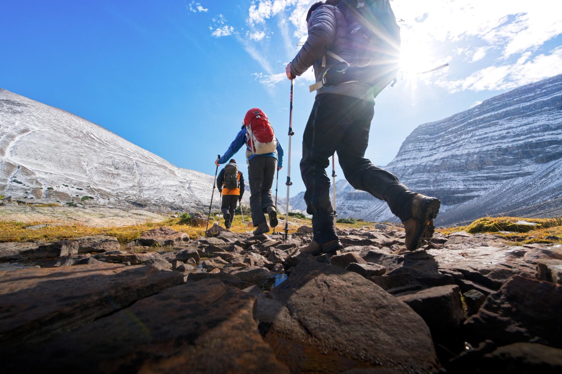 Three hikers with trekking poles walk along a rocky trail between steep mountain walls under a bright blue sky.