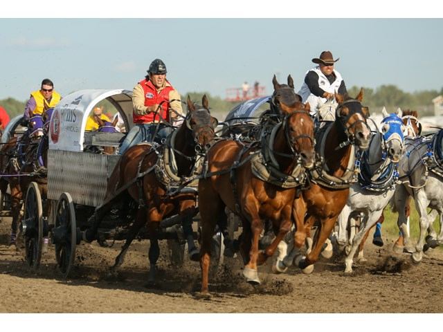 Chuckwagon teams race side by side as drivers urge their horses forward on a dirt track.