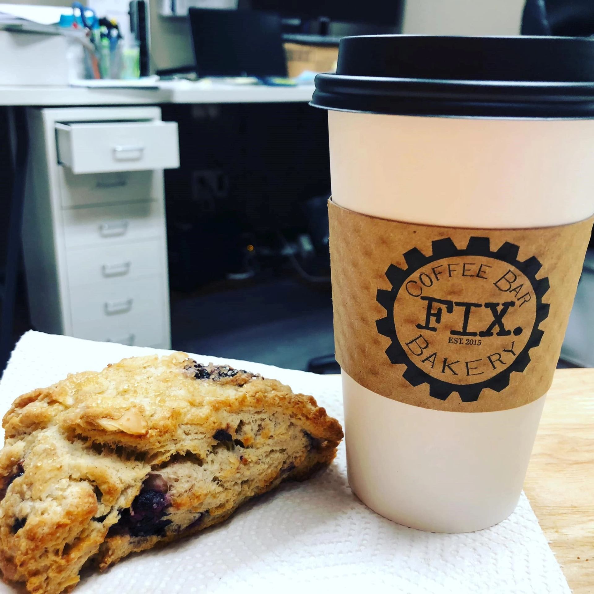 Coffee cup with FTX Coffee Bar Bakery sleeve beside scone on napkin in office.