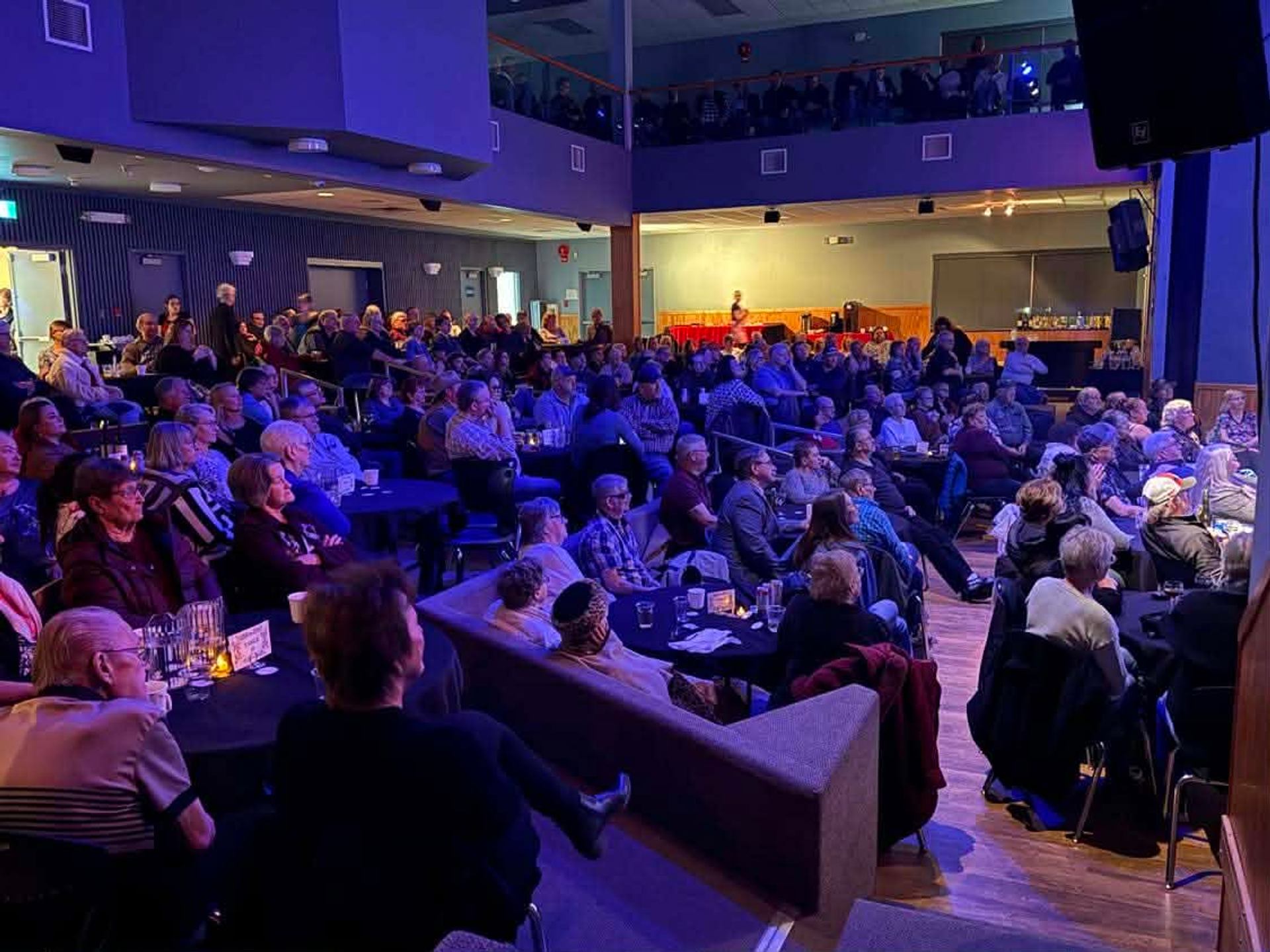 Audience members sit at tables in a crowded venue under blue lighting.