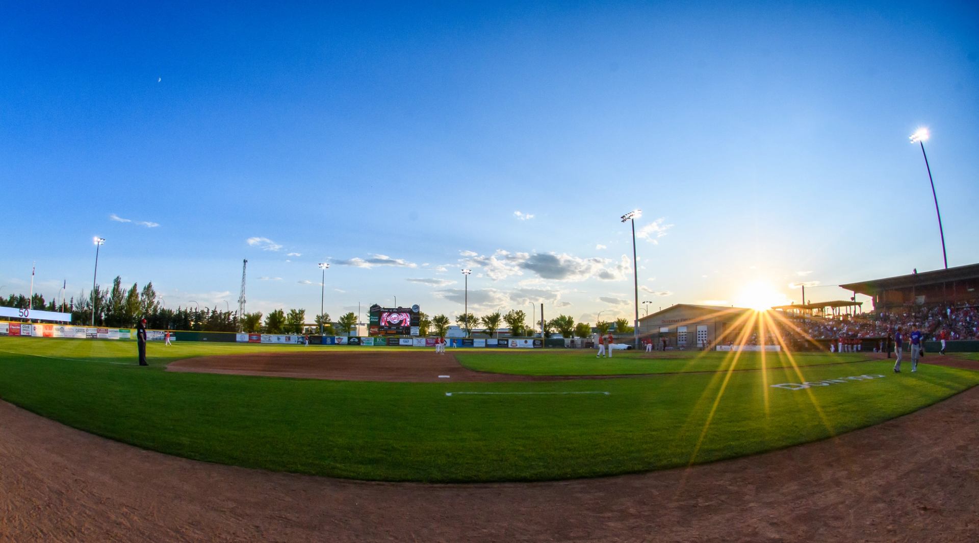 Baseball field at sunset with bright lights and clear blue sky.