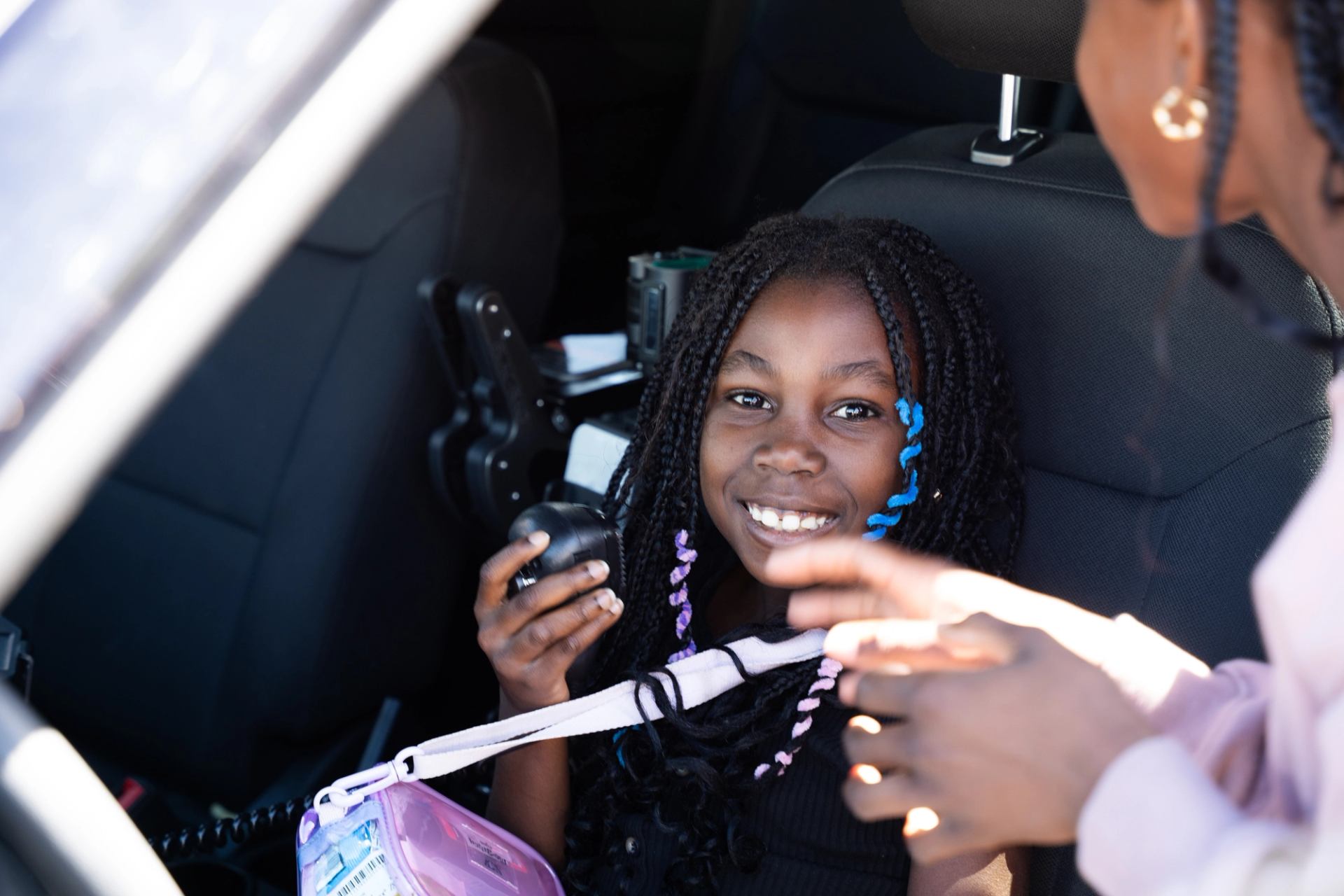 Smiling child holds a radio handset while seated beside an adult in a vehicle.