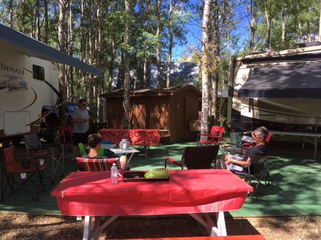Campsite picnic table with red tablecloth, chairs, and parked trailer among trees.