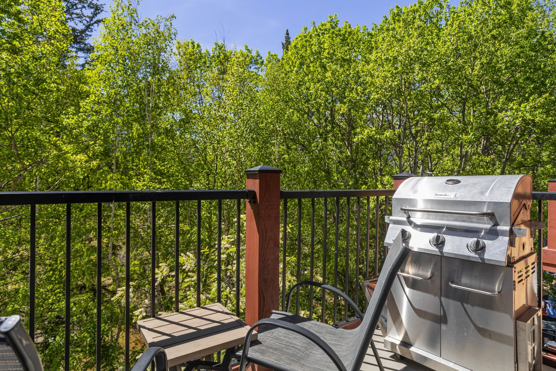 Balcony with stainless steel barbecue grill, chairs, and forest view in the background.
