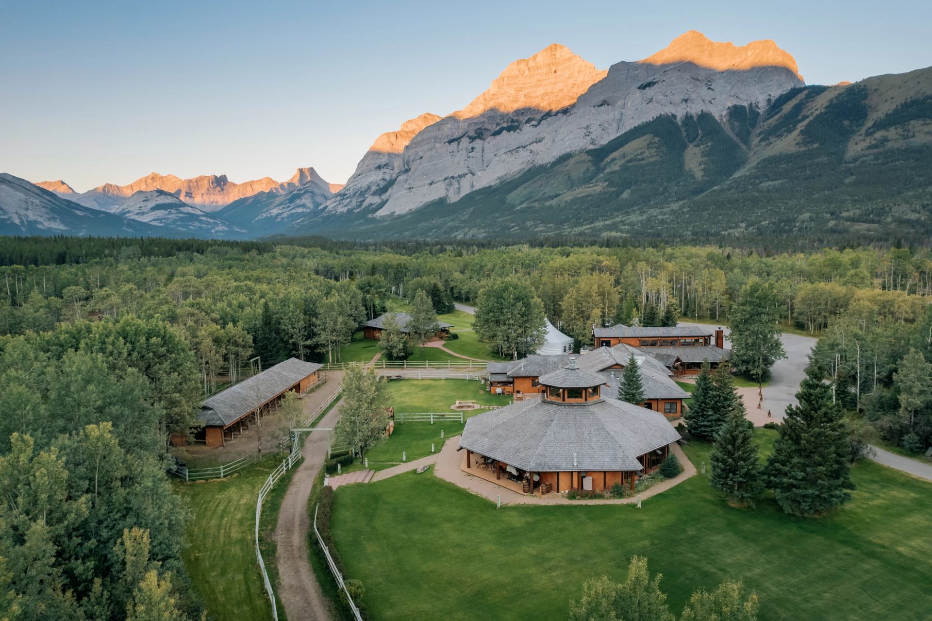 Aerial view of a rustic mountain lodge and stables surrounded by green forest, with sunlit mountains in the distance.