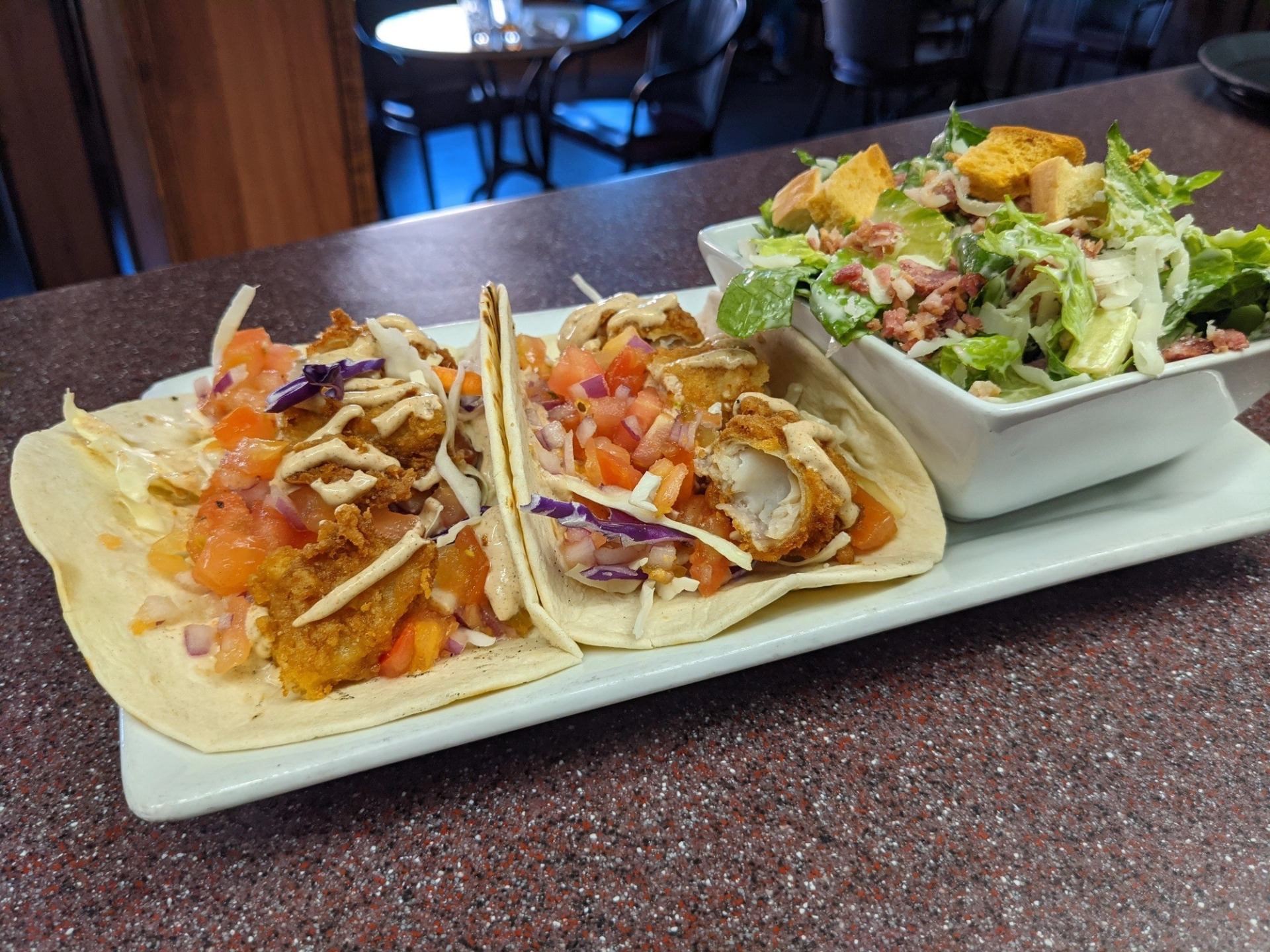 Plate with three fried fish tacos topped with vegetables beside a bowl of green salad.