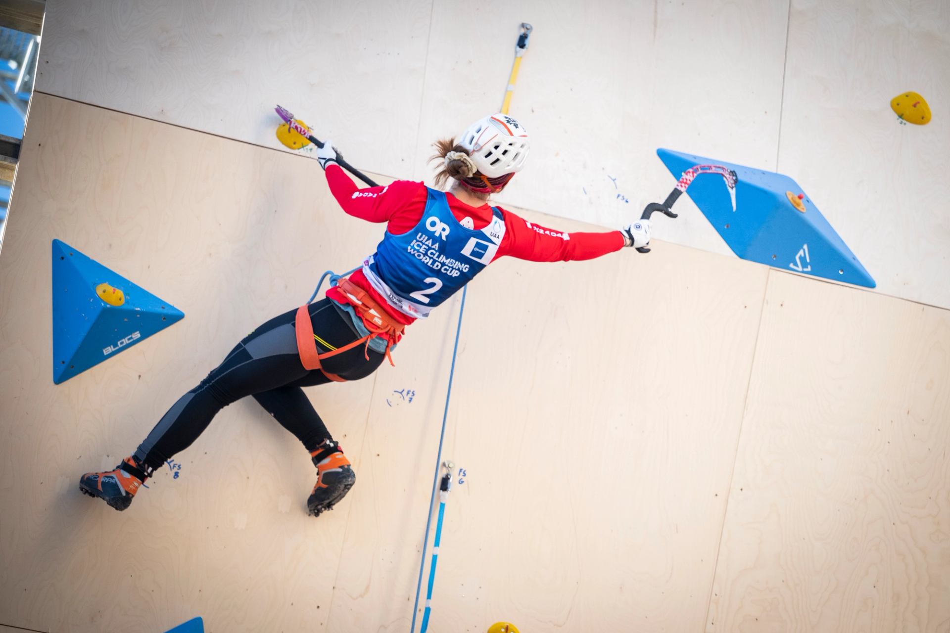 Athlete scaling an indoor wall using ice tools during a climbing event.