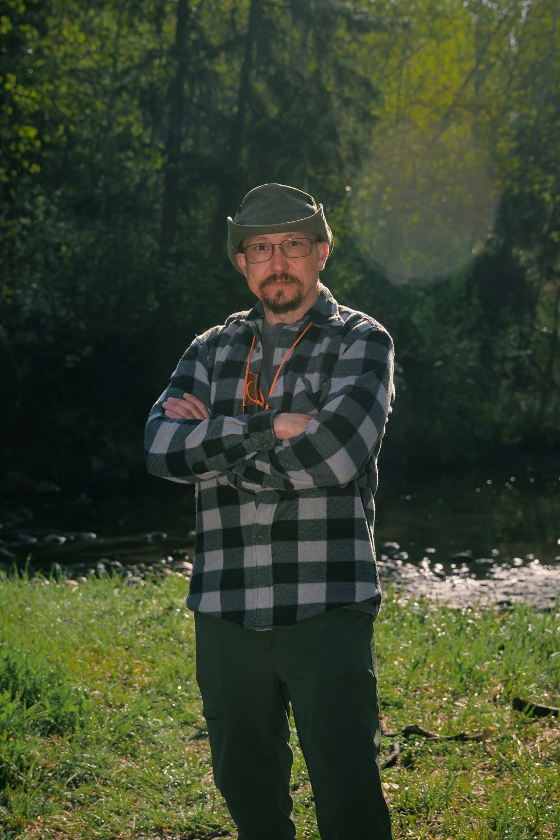 Person in plaid shirt and hat stands with crossed arms beside a forest stream.