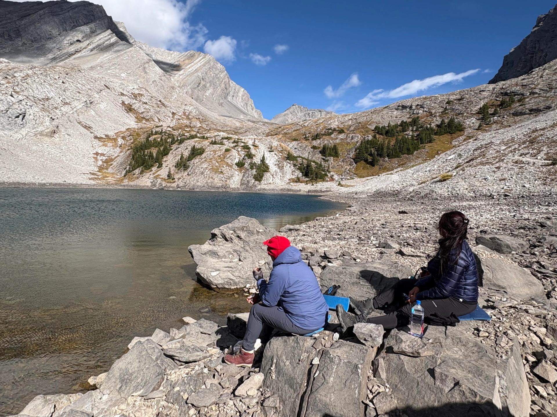Two hikers resting on rocky shore beside a clear alpine lake surrounded by rugged peaks.